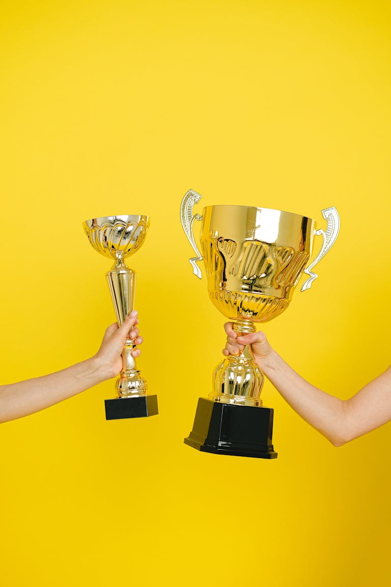 Two hands holding trophies against a vivid yellow backdrop, symbolizing success.